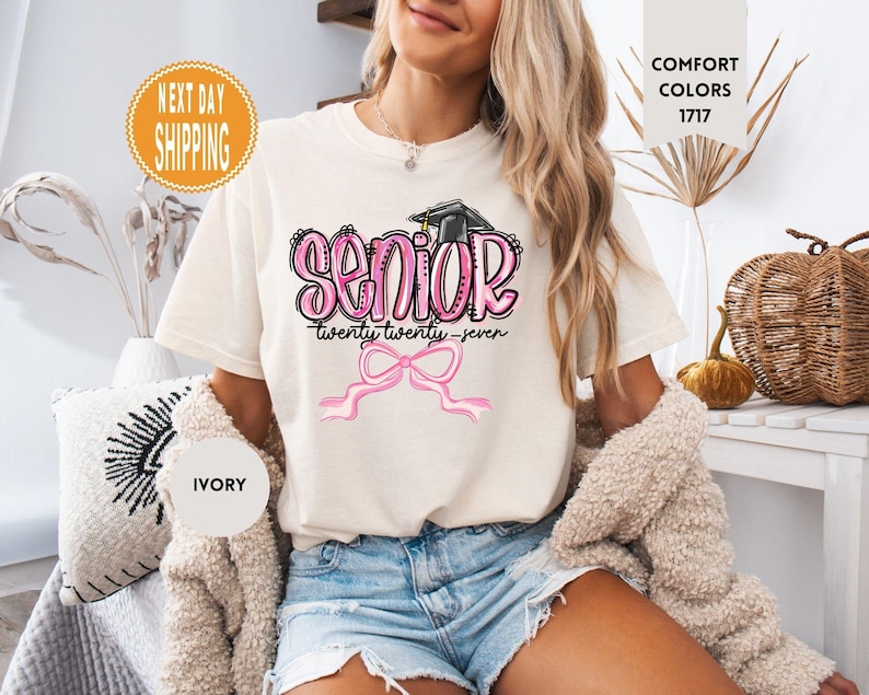 May include: An ivory short-sleeved t-shirt featuring a graphic with the word SENIOR in pink bubble letters, a black graduation cap, the text twenty twenty-seven, and a pink ribbon bow. The shirt includes a Comfort Colors 1717 label.