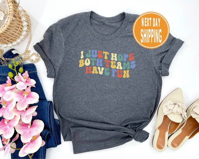 May include: A dark grey t-shirt with the text "I JUST HOPE BOTH TEAMS HAVE FUN" in rainbow colored letters.