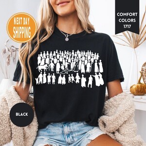 May include: Black t-shirt featuring a white graphic of a crowd of people in various poses. The shirt has the words "Comfort Colors 1717" printed on it. The shirt also has a "Next Day Shipping" sticker and a "Black" circle.