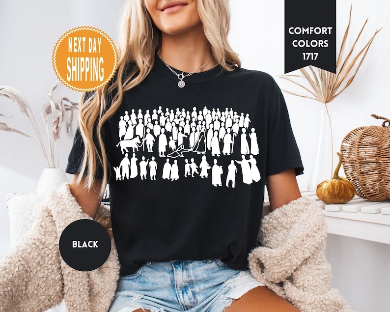 May include: Black t-shirt featuring a white graphic of a crowd of people in various poses. The shirt has the words "Comfort Colors 1717" printed on it. The shirt also has a "Next Day Shipping" sticker and a "Black" circle.