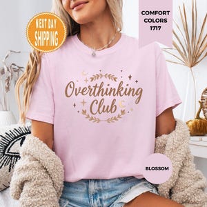 Puede incluir: Una camiseta rosa claro con la frase "Overthinking Club" en letras doradas, rodeada de un diseño floral. La camiseta es de la colección Comfort Colors 1717 y es de color Blossom.