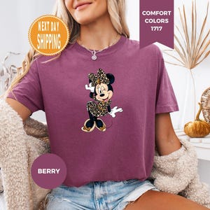 May include: Berry-colored t-shirt featuring a Minnie Mouse graphic in a leopard print outfit. The shirt has short sleeves and a crew neck. The image also includes text that says "Comfort Colors 1717" and "Berry".