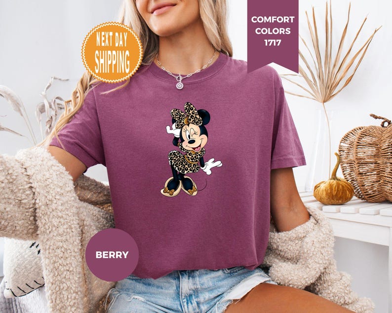 May include: Berry-colored t-shirt featuring a Minnie Mouse graphic in a leopard print outfit. The shirt has short sleeves and a crew neck. The image also includes text that says "Comfort Colors 1717" and "Berry".