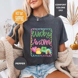 May include: Dark gray t-shirt with a colorful "Lucky Charm" graphic. The design includes a rainbow, heart, pot of gold, and a four-leaf clover. The shirt has a crew neck and short sleeves. The text "Comfort Colors 1717" is in the upper right corner.