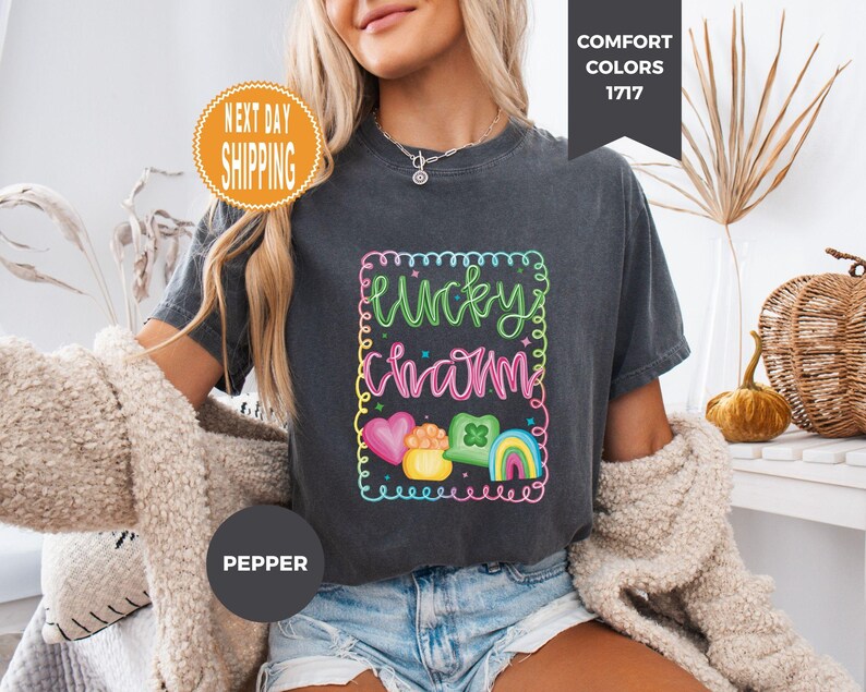 May include: Dark gray t-shirt with a colorful "Lucky Charm" graphic. The design includes a rainbow, heart, pot of gold, and a four-leaf clover. The shirt has a crew neck and short sleeves. The text "Comfort Colors 1717" is in the upper right corner.