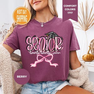 May include: A berry-colored short-sleeve t-shirt featuring a graphic print with the word SENIOR in large pink bubble letters, a black graduation cap, the text twenty twenty-seven, and a pink ribbon bow graphic centered below the text.