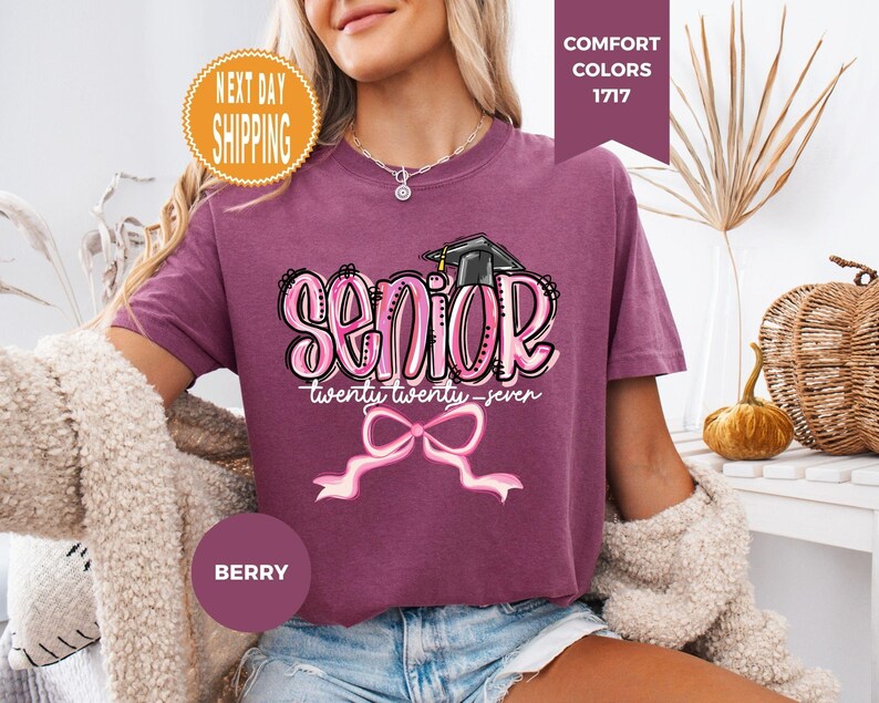 May include: A berry-colored short-sleeve t-shirt featuring a graphic print with the word SENIOR in large pink bubble letters, a black graduation cap, the text twenty twenty-seven, and a pink ribbon bow graphic centered below the text.
