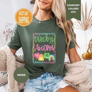 May include: Moss green t-shirt with a colorful "Lucky Charm" graphic. The design includes a rainbow, hearts, and a pot of gold. The shirt is a Comfort Colors brand.