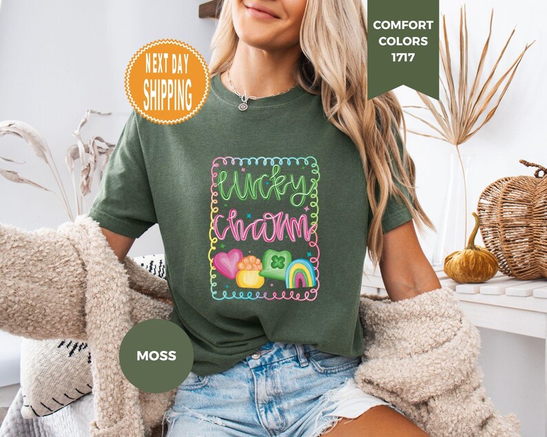 May include: Moss green t-shirt with a colorful "Lucky Charm" graphic. The design includes a rainbow, hearts, and a pot of gold. The shirt is a Comfort Colors brand.
