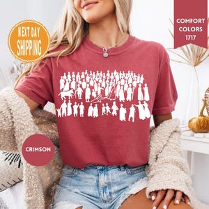 May include: Crimson-colored t-shirt featuring a white graphic of numerous figures. The shirt has short sleeves and is made of a soft material. The image also includes text that says "Comfort Colors 1717".
