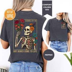 May include: Dark gray t-shirt featuring a skeleton holding a bouquet of colorful flowers. The design includes the text "Ezekiel 37:1-10" and "Dry Bones Come to Life". The shirt is a comfort color.