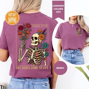 May include: Berry-colored t-shirt featuring a skeleton holding a bouquet of flowers. The skeleton is adorned with flowers and the text "Ezekiel 37:1-10" and "Dry Bones Come To Life" is printed on the shirt. The shirt is a Comfort Colors 1717.