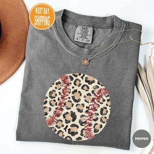 May include: A charcoal gray short-sleeve t-shirt featuring a circular graphic of a baseball with a tan and black leopard print pattern and red stitching. The shirt has a Comfort Colors brand label at the neck and the word Pepper printed in a circle.