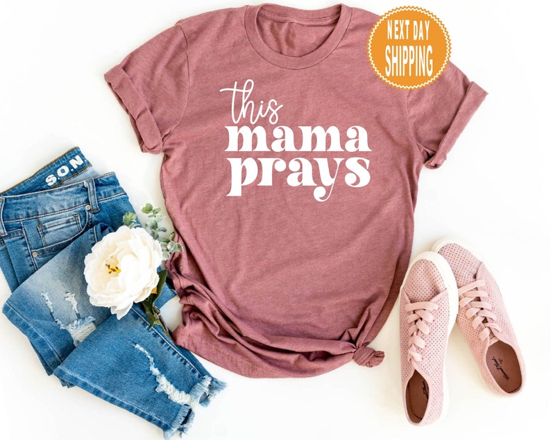 May include: A maroon t-shirt with the text "this mama prays" in white lettering. The text is in a retro font style.