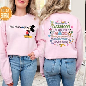 May include: Pink sweatshirt with a Mickey Mouse design on the front and a Disney-themed classroom quote on the back. The quote reads "Welcome to our classroom where the magic never stops and the adventure never ends!"