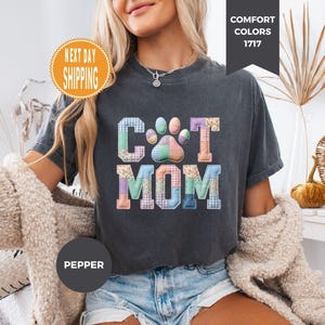 May include: Dark gray t-shirt with the words "Cat Mom" in a patchwork design. The letter "C" is in blue and green plaid, the "A" is in pink and purple, and the "T" is in orange. The paw print is in pastel colors.