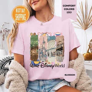 May include: A light pink t-shirt with a colorful graphic of Walt Disney World. The graphic includes images of the park's attractions and the words "Walt Disney World". The shirt also has the words "Comfort Colors 1717" and "Blossom".