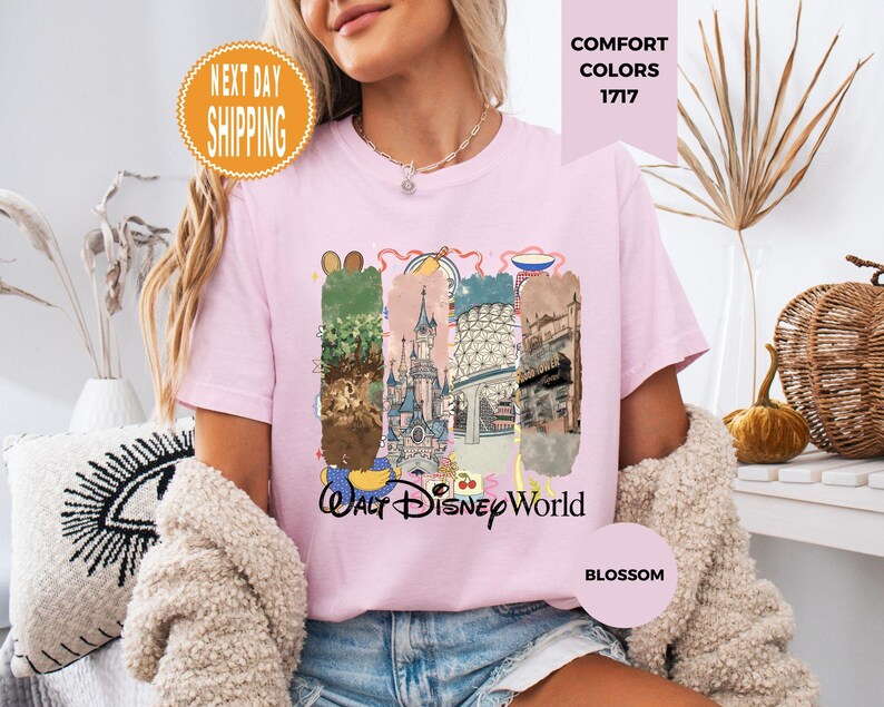May include: A light pink t-shirt with a colorful graphic of Walt Disney World. The graphic includes images of the park's attractions and the words "Walt Disney World". The shirt also has the words "Comfort Colors 1717" and "Blossom".
