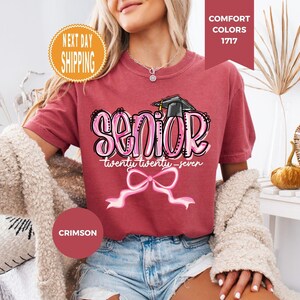 May include: A crimson short-sleeve t-shirt featuring a graphic print with the word SENIOR in large pink letters, the text twenty twenty-seven, a black graduation cap, and a pink ribbon bow. The shirt has a crew neckline and a relaxed fit.