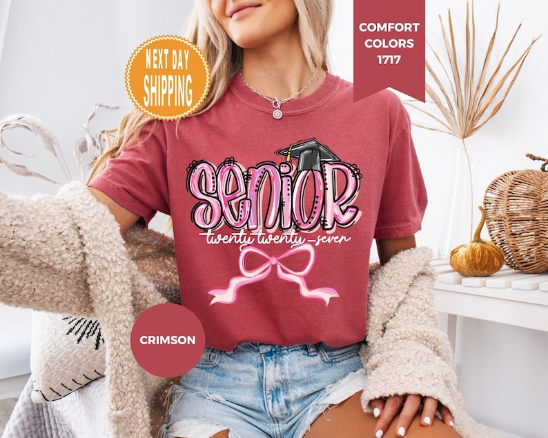 May include: A crimson short-sleeve t-shirt featuring a graphic print with the word SENIOR in large pink letters, the text twenty twenty-seven, a black graduation cap, and a pink ribbon bow. The shirt has a crew neckline and a relaxed fit.
