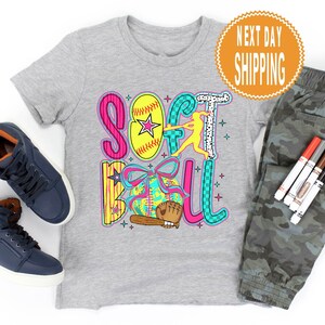 May include: A gray t-shirt with a colorful "Softball" graphic, a softball, and a batter. The image includes navy blue high-top sneakers, camouflage pants, and markers. A round orange sticker says "Next Day Shipping".