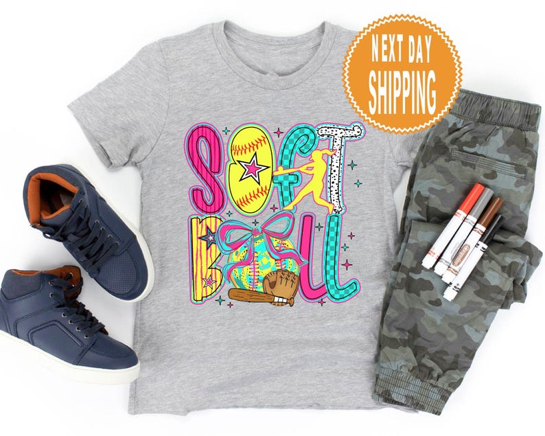 May include: A gray t-shirt with a colorful "Softball" graphic, a softball, and a batter. The image includes navy blue high-top sneakers, camouflage pants, and markers. A round orange sticker says "Next Day Shipping".