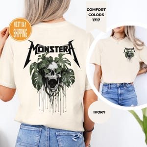 Puede incluir: Camiseta de color marfil con un diseño de calavera y la palabra "MONSTERA" en negro, rodeada de hojas verdes. La camiseta tiene cuello redondo y mangas cortas. También se ve una versión más pequeña del diseño en la parte delantera.