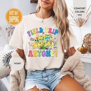Op de afbeelding: Een wit T-shirt met een kleurrijke afbeelding van een schoolbus en de tekst "Field Trip Anyone?" erop gedrukt. Het shirt wordt gedragen door een persoon met lang blond haar.