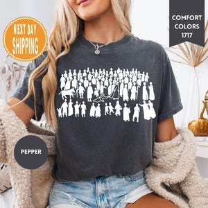 May include: Dark gray t-shirt with a white graphic of numerous silhouetted figures. The shirt has the text "COMFORT COLORS 1717" and "PEPPER" on it. The shirt is a crew neck and short-sleeved.