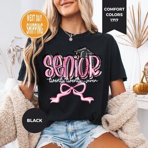 May include: A black t-shirt featuring a graphic with the word SENIOR in pink bubble letters, a black graduation cap, the text twenty twenty-seven, and a pink ribbon bow. The design is centered on the chest of the short-sleeved garment.