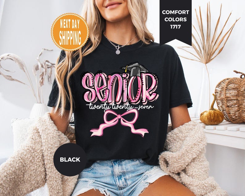 May include: A black t-shirt featuring a graphic with the word SENIOR in pink bubble letters, a black graduation cap, the text twenty twenty-seven, and a pink ribbon bow. The design is centered on the chest of the short-sleeved garment.
