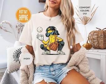 May include: An ivory t-shirt featuring a graphic of a yellow banana character wearing a black baseball cap, blue sunglasses, and headphones. The banana is in a catcher's stance with a brown baseball glove, knee pads, and a baseball nearby.