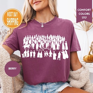 May include: Berry-colored t-shirt with a white graphic of numerous figures. The shirt has a crew neck and short sleeves. The words "Comfort Colors 1717" are in a burgundy rectangle. A round sticker says "Next Day Shipping".