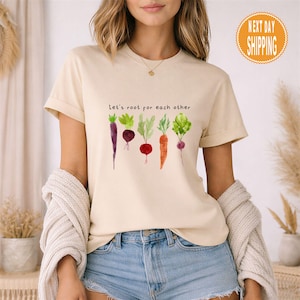 May include: A beige t-shirt featuring the phrase "Let's root for each other" and a watercolour illustration of root vegetables. The vegetables include purple carrots, beetroot, and carrots. The shirt has short sleeves.