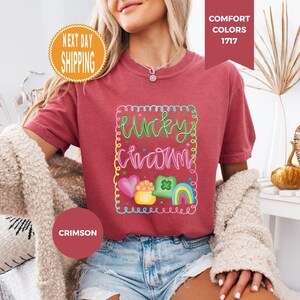 May include: Crimson-colored t-shirt with the words "Lucky Charm" in a colorful, whimsical design. The design includes a rainbow, hearts, and a pot of gold. The shirt is a Comfort Colors 1717 style.