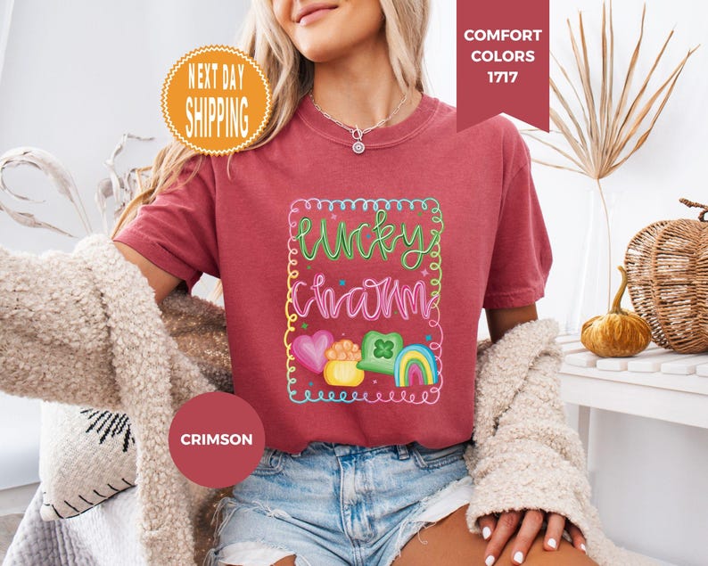 May include: Crimson-colored t-shirt with the words "Lucky Charm" in a colorful, whimsical design. The design includes a rainbow, hearts, and a pot of gold. The shirt is a Comfort Colors 1717 style.