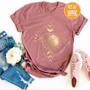 Op de afbeelding: Een mauve t-shirt met een gouden zon- en maandesign. Het ontwerp bevat een zon met een bloem erin en een wassende maan met sterren. De tekst "NEXT DAY SHIPPING" staat op een gele cirkel rechtsboven.