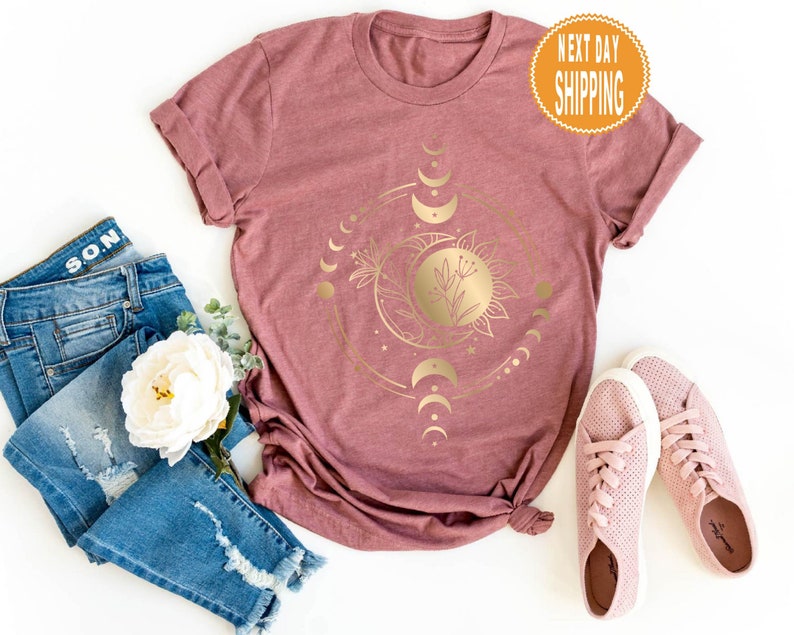 Op de afbeelding: Een mauve t-shirt met een gouden zon- en maandesign. Het ontwerp bevat een zon met een bloem erin en een wassende maan met sterren. De tekst "NEXT DAY SHIPPING" staat op een gele cirkel rechtsboven.