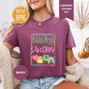 May include: Berry-colored t-shirt with a colorful "Lucky Charm" graphic. The graphic includes a rainbow, shamrock, and heart. The shirt has a crew neck and short sleeves. The words "Next Day Shipping" are in a circular orange badge.