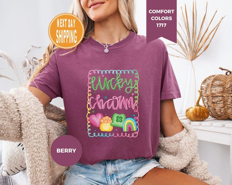 May include: Berry-colored t-shirt with a colorful "Lucky Charm" graphic. The graphic includes a rainbow, shamrock, and heart. The shirt has a crew neck and short sleeves. The words "Next Day Shipping" are in a circular orange badge.