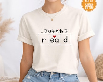 Może przedstawiać: Kremowy t-shirt z czarnym napisem "I teach kids to read", z czerwonym sercem zastępującym "a" w słowie "read". Koszulka ma krótkie rękawy i jest wykonana z miękkiego materiału.