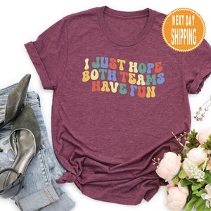 May include: Burgundy t-shirt with the text "I just hope both teams have fun" in rainbow colors.