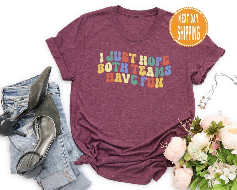 May include: Burgundy t-shirt with the text "I just hope both teams have fun" in rainbow colors.
