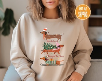 May include: Beige sweatshirt featuring three dachshunds in festive attire. One wears a Santa hat, another carries a Christmas tree, and the third is on skis. The text "Dachshund Through The Snow" is also visible.