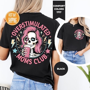 May include: Black t-shirt with a graphic featuring a skull with pink hair drinking from a pink cup, surrounded by the words "OVERSTIMULATED MOMS CLUB". The design includes lightning bolts and stars.