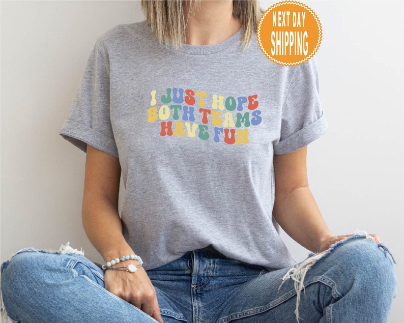 May include: Gray t-shirt with the text "I just hope both teams have fun" in rainbow colors.