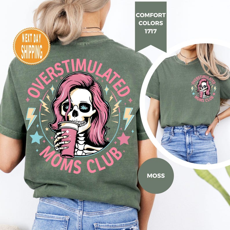 May include: Moss green t-shirt with a graphic on the back featuring a skull with pink hair holding a pink coffee cup, surrounded by text that reads "OVERSTIMULATED MOMS CLUB." The front of the shirt has a smaller version of the graphic. The shirt is made of a soft, comfortable material.