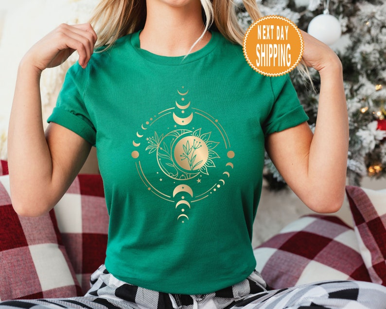 Op de afbeelding: Een groen T-shirt met een gouden ontwerp van een zon en maan met bloemmotieven. Het ontwerp is omringd door een cirkel van kleine manen.