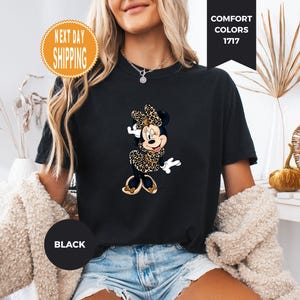 May include: Black t-shirt featuring a Minnie Mouse graphic in a leopard print design. The shirt has the text "Comfort Colors 1717" and "Black". The shirt is worn by a person.