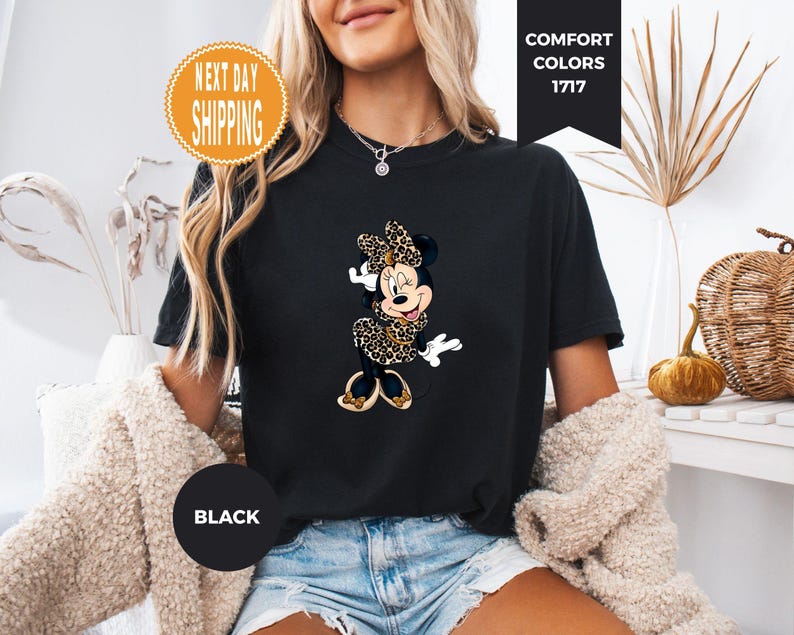 May include: Black t-shirt featuring a Minnie Mouse graphic in a leopard print design. The shirt has the text "Comfort Colors 1717" and "Black". The shirt is worn by a person.